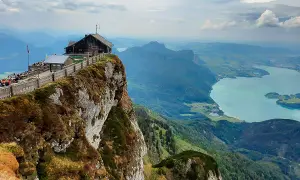 Ein Gasthaus thront am Felsvorsprung, die Himmelspforte am Schafberg, mit Blick auf einen türkisfarbenen See und grüne Berge.