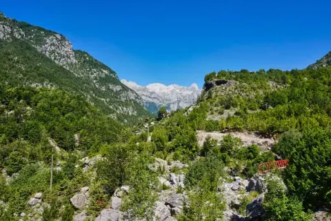 Grüne Hänge und felsige Gipfel in den albanischen Alpen unter strahlend blauem Himmel. Ein kleiner roter Steg führt über einen steinigen Bachlauf.