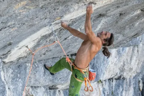 Alexander Huber klettert an einer steilen Felswand, sein Körper angespannt, die Hände greifen nach kleinen Vorsprüngen.