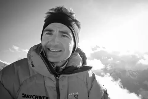 Ueli Steck, lächelnd in warmer Bergkleidung, blickt in die sonnige, wolkenverhangene Bergwelt.