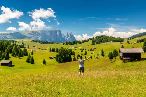 Ein Wanderer genießt die Aussicht auf eine Alm mit Berghütten und Wäldern. Perfekt für eine Mehrtageswanderung in den Dolomiten.