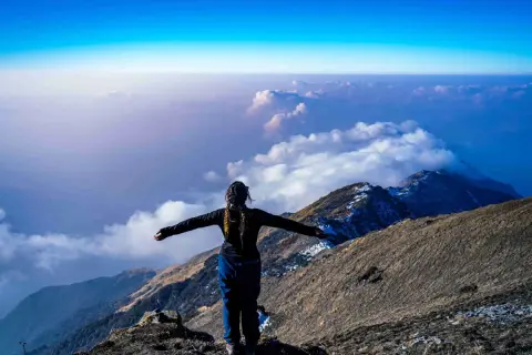 Eine Frau steht auf einem Berggipfel, die Arme ausgebreitet, und blickt auf eine Wolkendecke. Die Frage "wie hoch ist der Kilimandscharo?" hallt in der Weite wider.