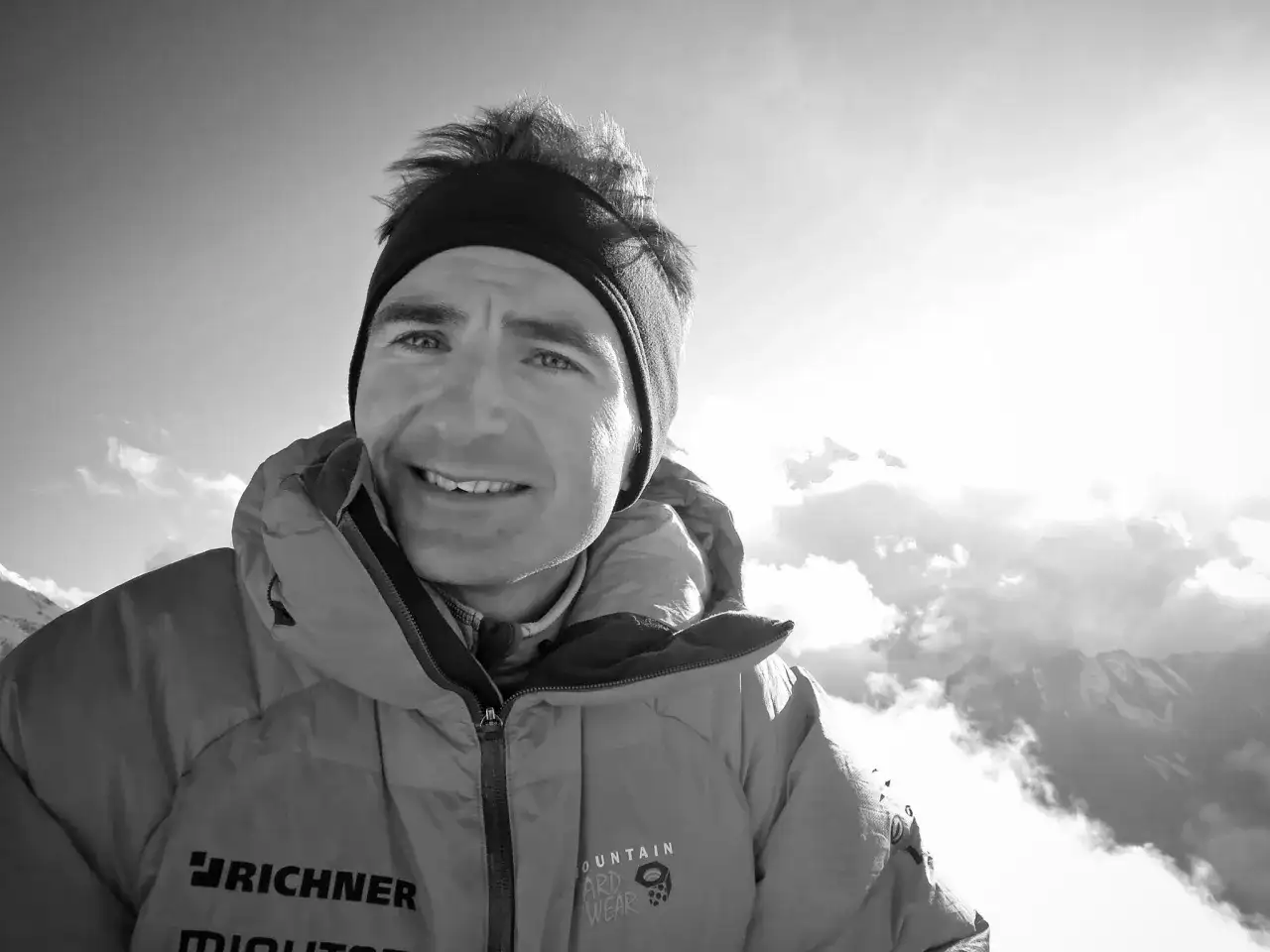 Ueli Steck, lächelnd in warmer Bergkleidung, blickt in die sonnige, wolkenverhangene Bergwelt.
