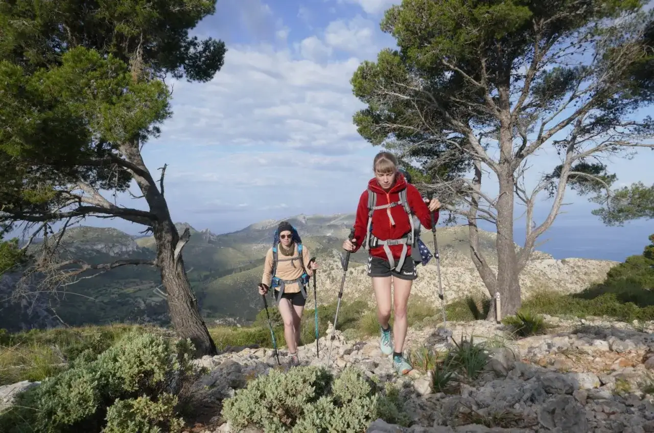 Zwei Wanderinnen mit Rucksäcken und Stöcken auf einem felsigen Pfad, umgeben von Bäumen und mit Blick auf eine hügelige Landschaft und das Meer. Eine tolle Tour, wie auf komoot geplant.