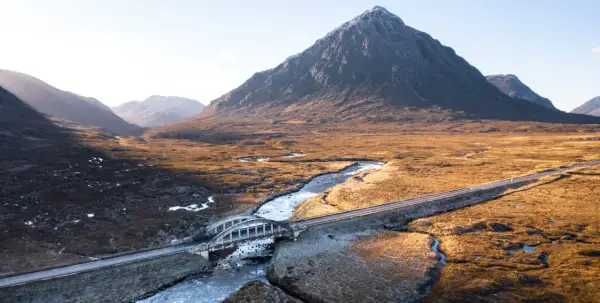 Majestätische Berge der Schottland Highlands, ein Fluss schlängelt sich durch das goldene Gras, überquert von einer Brücke.