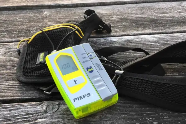 Ein Pieps lawinen sender mit Display und Suchfunktion, daneben eine schwarze Tasche mit gelben Akzenten, liegt auf einer Holzoberfläche.